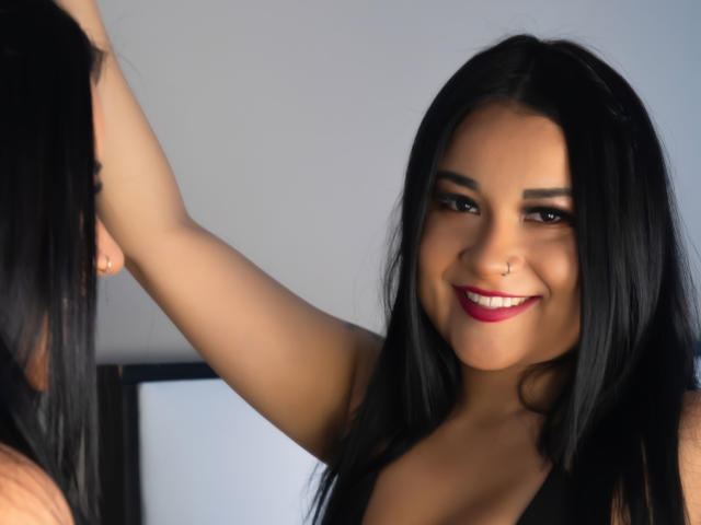 Chloe Rouse, met zwart haar, lachend naar haar spiegelbeeld, arm omhoog, met een neusring, rode lippenstift en een schattig gezicht, gekleed in een zwarte top, in een spiegel-selfie..