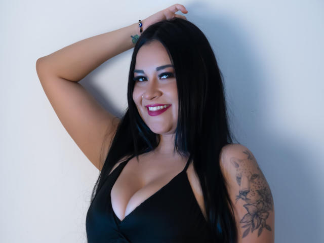 Chloe Rouse, con cabello negro, sonriendo, vestida con una blusa negra, con el brazo levantado, mostrando tatuajes en su hombro derecho, de busto medio, con lápiz labial rosa, maquillaje de ojos azul, y un fondo de pared blanca..