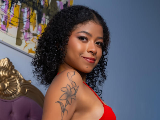 SaraTonson with curly black hair, brown skin, red top, nose ring, and floral tattoo on her shoulder, smiling at the camera. Boobs visible, blue wall background.