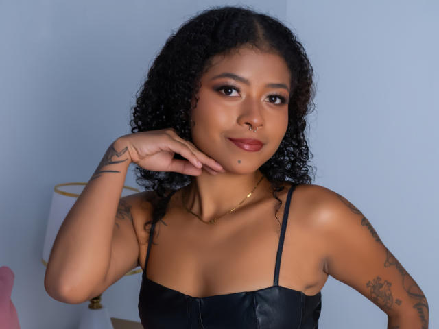 SaraTonson with curly black hair, tan skin, nose ring, and tattoos, wearing a black leather top, touching her chin, smiling with red lipstick. Her tits are small.