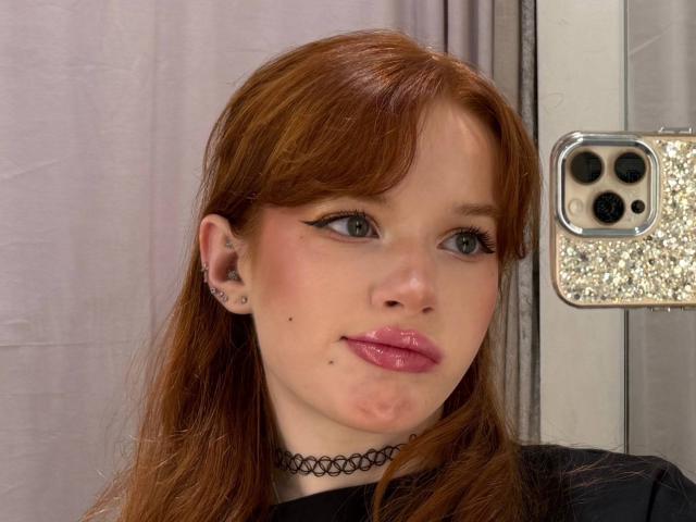Margo Tamminen, avec des cheveux roux, des yeux bleus, des lèvres roses et brillantes, un collier noir, un petit nez, des piercings aux oreilles, tenant un téléphone scintillant, une petite imperfection sur le menton, portant un haut noir..