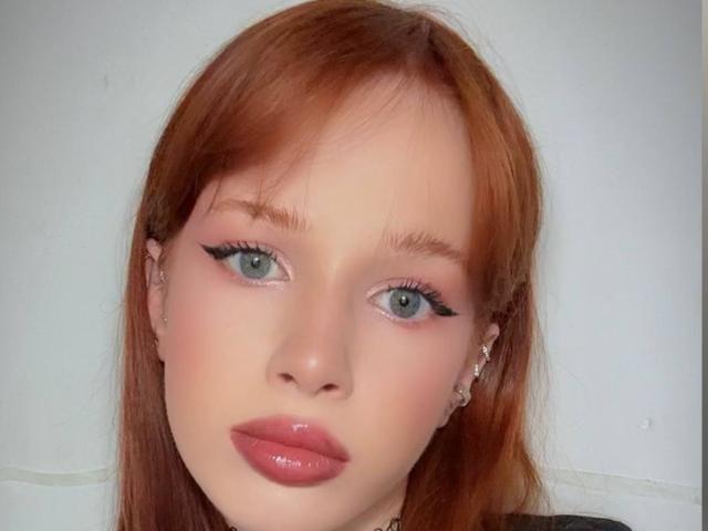 El rostro de MargoTamminen, con piel pálida, cabello rojo, ojos azules, delineador de ojos negro, labios rosados brillantes y pequeños aretes de plata, mirando directamente a la cámara..