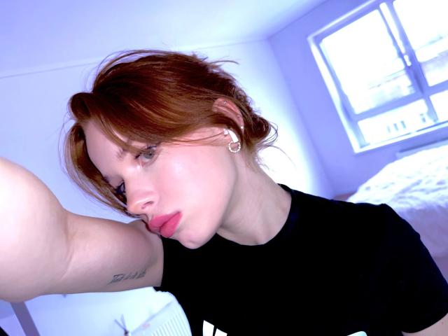 Margo Tamminen, met rood haar, een zwart shirt en een neusring, met een frons op haar lippen, met een arm omhoog waarbij een tatoeage zichtbaar is, in een kamer met blauw licht..