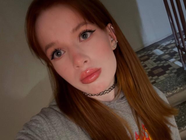 Margo Tamminen, met rood haar, blauwe ogen, glanzende roze lippen en een zwarte choker, draagt een grijze hoodie en kijkt met een lichte frons naar de camera..