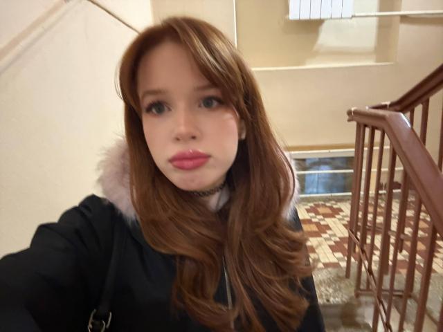 Margo Tamminen tiene el pelo largo y castaño, lápiz labial rosa y un abrigo negro con cuello de piel. Está de pie en una escalera con barandilla de madera. Sus glúteos no son visibles..