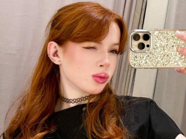 MargoTamminen, con i capelli rossi, che fa l'occhiolino, indossa una collana nera, tiene in mano un telefono scintillante, labbra rosa, maglietta nera, piccolo piercing all'orecchio, selfie allo specchio..