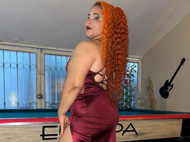 NightPasssion with orange curly hair in a tight maroon dress, showing her ass, standing by a pool table, looking back with dark red lipstick.