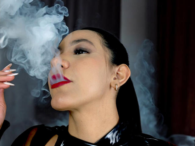 ThaleiaStone with black hair, red lipstick, and long nails, inhaling smoke from her hand, wearing a shiny black top, with a small hoop earring in her left ear.
