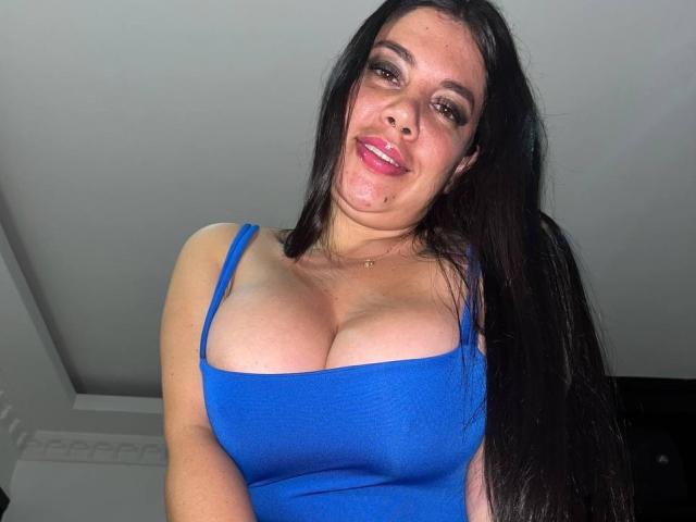AndreaBusty with long black hair, blue tank top, showing her large tits, smiling with pink lipstick, nose piercing, and a small beauty mark on her right cheek.