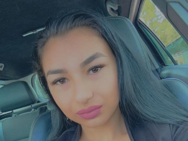 CataleyaStarr hat eine mittelbraune Haut, langes schwarzes Haar, dicke Augenbrauen und pinken Lippenstift. Sie sitzt in einem Auto mit schwarzen Ledersitzen. Ihre braunen Augen schauen in die Kamera..