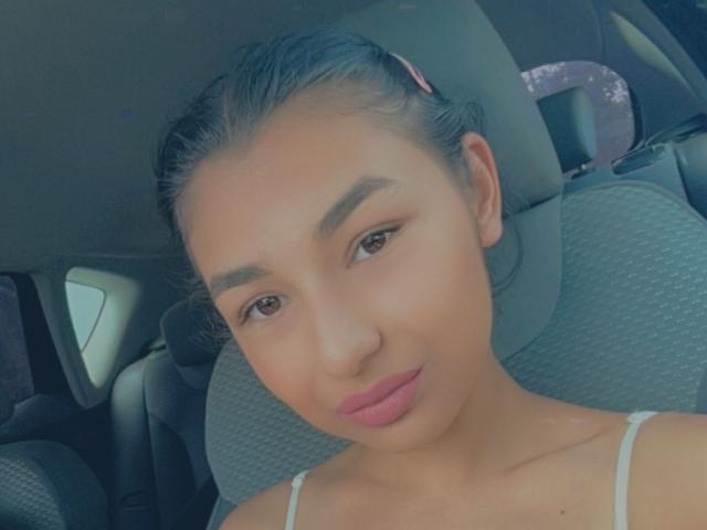 CataleyaStarr with smooth brown skin, pink lipstick, and brown eyes, wearing a white spaghetti strap top, seated in a car with grey upholstery.