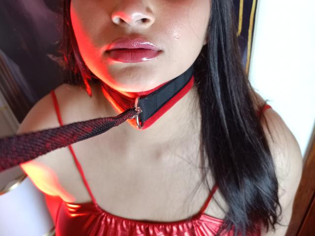 GraceBlue69's face, glossy pink lips, black mesh collar with red trim, and leash in mouth, wearing red satin top, dark straight hair, red light on skin.