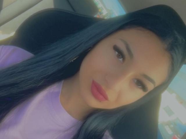 CataleyaStarr, con lunghi capelli neri, una maglietta viola, rossetto rosso e occhi marroni, seduta in un'auto, che guarda la fotocamera, mostrando il suo viso e la parte superiore del corpo..