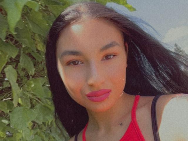 CataleyaStarr, avec ses longs cheveux noirs, ses yeux marrons, son rouge à lèvres rouge et un petit tatouage sur le cou, portant un haut rouge, debout devant des feuilles vertes, souriant à l'appareil photo..