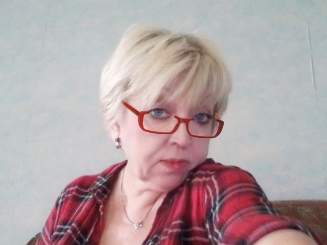 AnjelElinor with short blonde hair, red glasses, and a red plaid shirt, has a necklace and a serious expression. Boobs visible under the shirt.