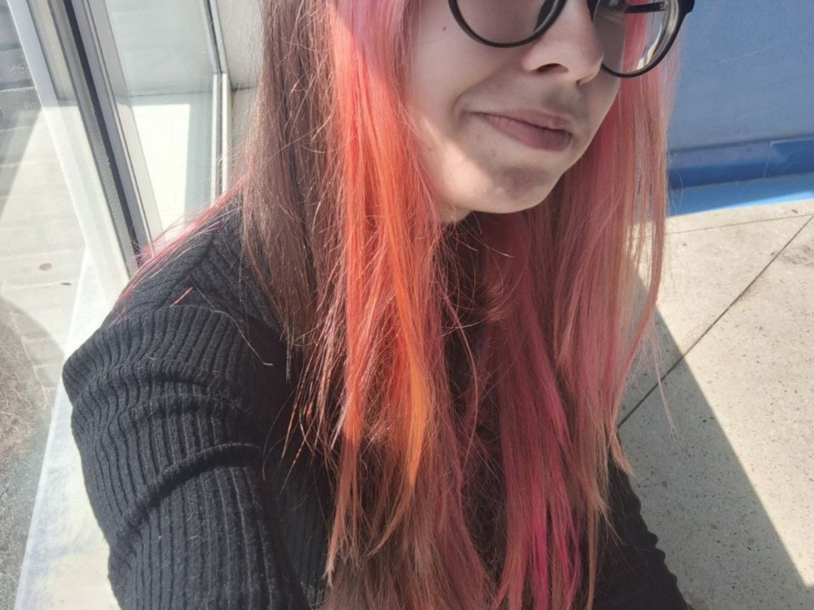 DorotheaMcmilliam tem o cabelo tingido de rosa e laranja, veste uma camiseta preta texturizada, usa óculos e sorri levemente, mostrando o nariz e os lábios. Ela está ao ar livre, sob a luz do sol..
