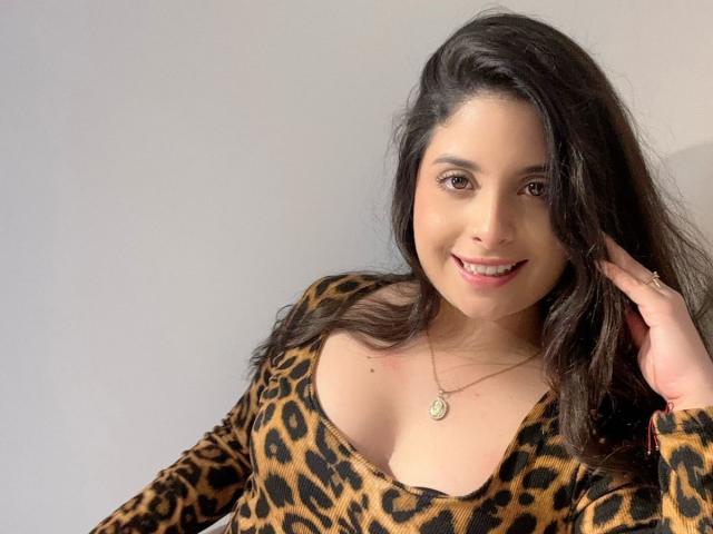 Luciana Blake, con cabello oscuro y ondulado, sonriendo, vistiendo una blusa de estampado de leopardo, mostrando sus senos, y un collar de oro con un dije en forma de corazón, tocándose el cabello..