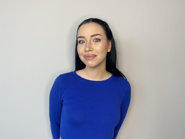 TiaParsell, con cabello negro largo, ojos azules y lápiz labial rosa, vestida con una camiseta azul ajustada de manga larga, que muestra sus pequeños senos firmes. Está sonriendo suavemente a la cámara..