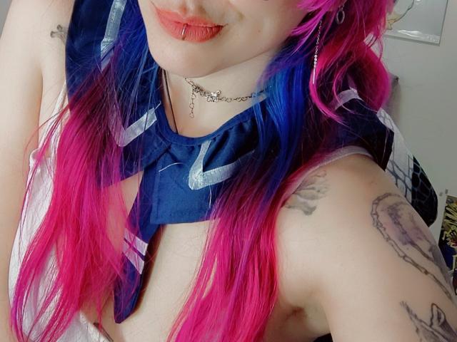 NatashaLau mit leuchtend rosa und blauen Haaren, die ein blaues Hemd mit weißen Streifen trägt und dabei ihre Brust und Tattoos zeigt, hat roten Lippenstift und eine silberne Kette um den Hals..