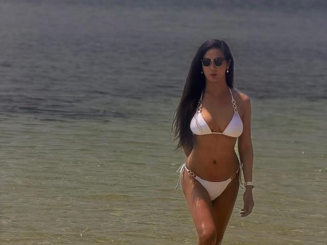 TopLadyManForU mit langen, schwarzen Haaren, die ein weißes Bikini trägt, hat kleine Brüste, gebräunte Haut und geht in flachem Wasser, wobei sie eine Sonnenbrille trägt..