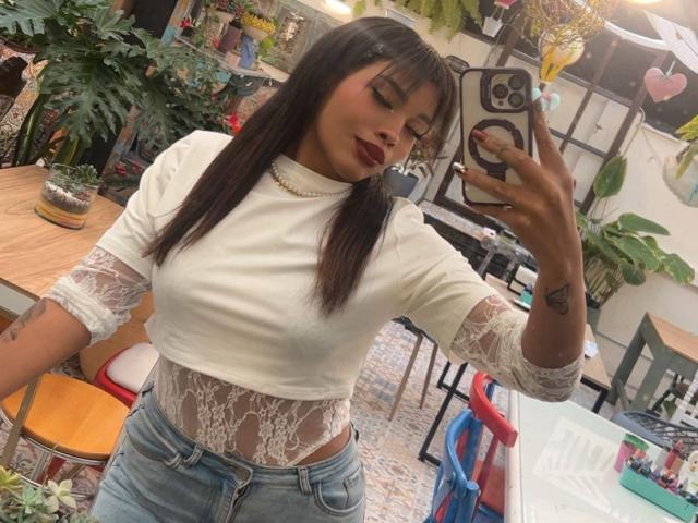 LexieBloom with dark hair, wearing a white lace top, showing her tits through the lace, and blue jeans, taking a mirror selfie, holding a phone with a ring case.