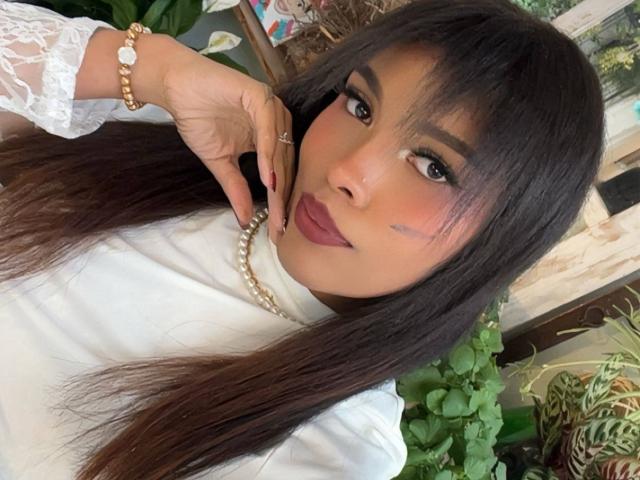 Lexie Bloom, con cabello largo y oscuro, vestida con una blusa blanca de encaje, un collar de perlas y lápiz labial rosa, tiene un pequeño moretón debajo del ojo y una expresión linda y segura..
