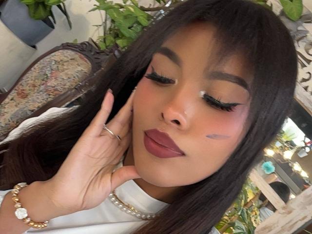 LexieBloom, con cabello negro largo, piel morena, lápiz labial rojo oscuro, sombra de ojos morada y pestañas negras, tocándose la cara, vistiendo una blusa blanca, una pulsera dorada y un collar de perlas..