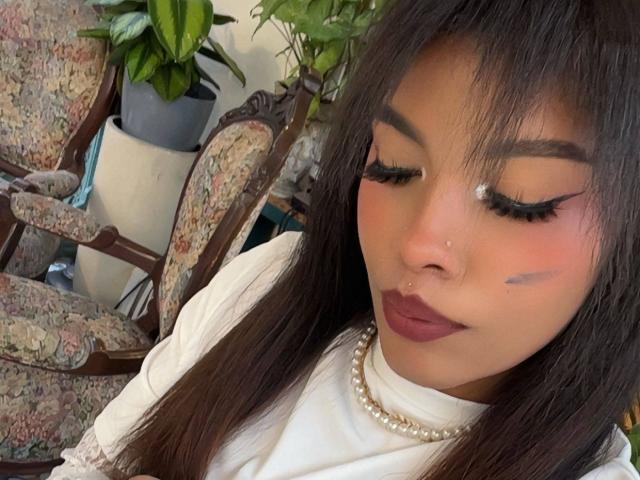 LexieBloom with dark hair, wearing white, pearl necklace, bold eyeliner, dark lipstick, nose ring, and subtle blue makeup on cheek. Floral-patterned chair and potted plant in background.
