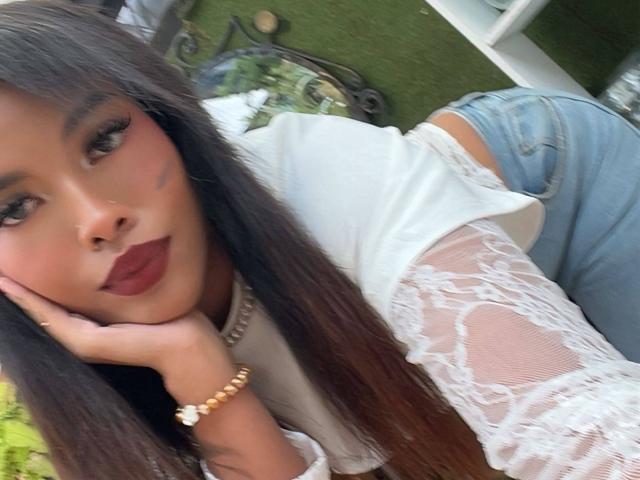 LexieBloom with long dark hair, wearing a white lace top, blue jeans, and a gold bracelet, resting her head on her hand, showing off her full, pouty ass.