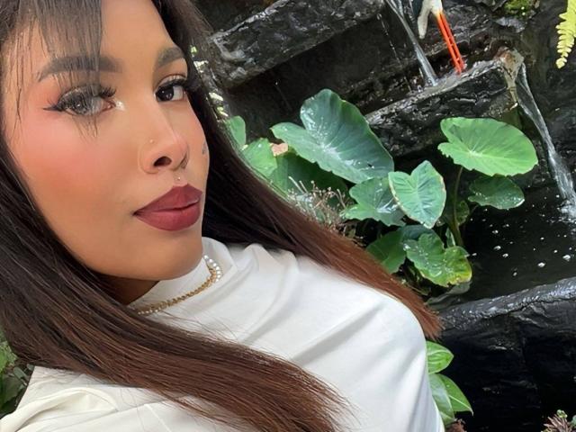 LexieBloom with dark hair, red lipstick, and a nose ring, wearing a white top and pearl necklace, standing next to a waterfall with green leaves.