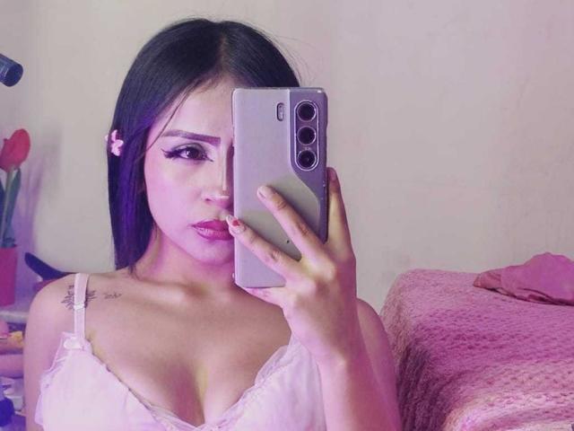 KimiNaru with dark hair, pink eyeliner, and white lace top, holding a silver phone, showing her boobs and a small tattoo on her right shoulder. Pink bed in background.