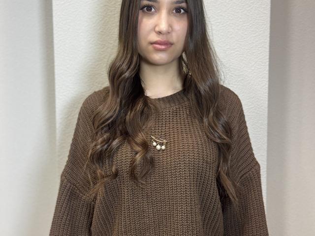 ShawneeLinderholm with long, wavy brown hair, wearing a brown knitted sweater, and a small butterfly necklace, standing against a white textured wall. Boobs, brown eyes, and pink lips.
