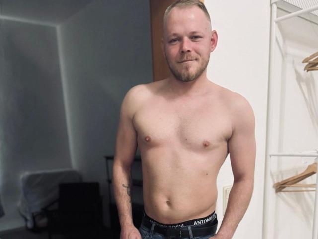 Colehunter, torse nu, avec un léger sourire, des cheveux blonds courts, une barbe légère et un tatouage sur son bras droit, portant des sous-vêtements noirs Antoroni, se tient dans une pièce..