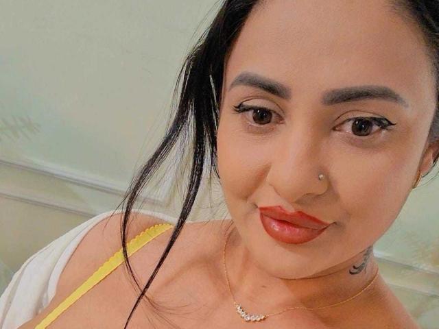 MayaMusee has dark eyes, red lipstick, black hair in a ponytail, a nose ring, and a necklace with a small diamond, showing yellow straps on her tits.