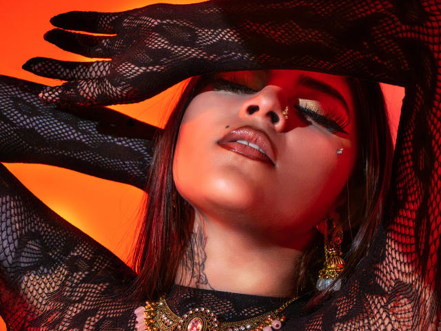 CoriPaige with her eyes closed, wearing black lace gloves, a nose piercing, and a necklace, her lips slightly parted, a small tattoo on her neck, against a red background.