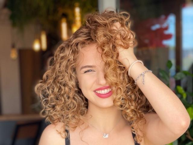 JacqueHailstone, met krullend blond haar, lacht met rode lippenstift, heeft haar hand in haar haar, draagt een zwarte top, een zilveren ketting en armband, heeft een lichte huid en ziet er schattig en aantrekkelijk uit..