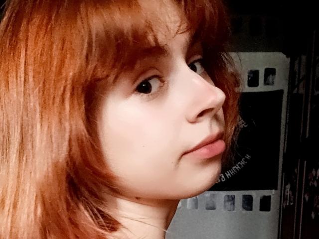 MireilleSpotwood has red hair, pale skin, and is looking to the side with a slight pout. Her small nose and subtle makeup are visible. Her eyes are dark.