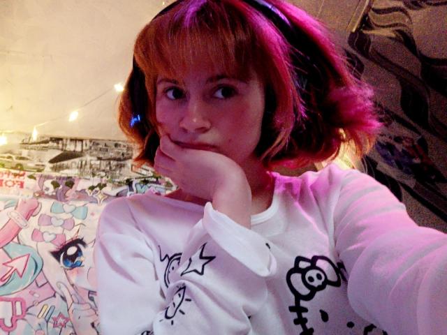 MireilleSpotwood with pink-tinted red hair, wearing a white shirt with black floral prints, head resting on hand, smiling, wearing black headphones, in a softly lit room.
