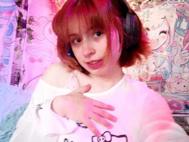 MireilleSpotwood with pink-tinged brown hair, wearing headphones, a white off-shoulder shirt, and a cute smile, touching her chest with her hand.