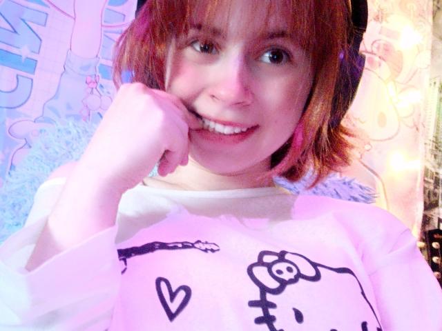 MireilleSpotwood with short red hair, smiling, biting her finger, wearing a white shirt with a cute black doodle, pink and purple light. Her tits are not visible.