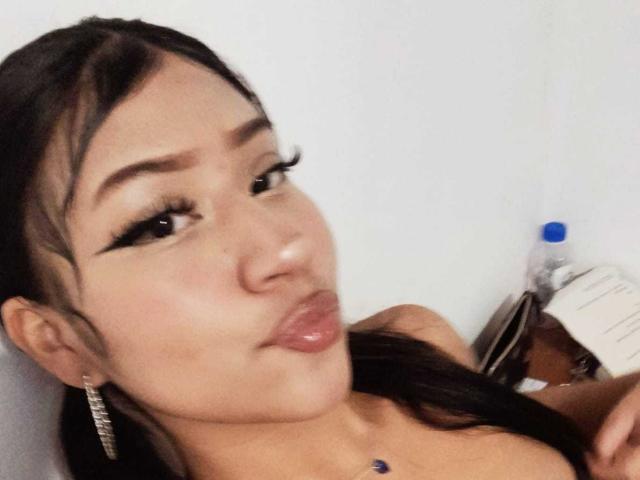 BonnieBond with pouty lips, long black eyelashes, and dark brown hair in a side ponytail, wearing a necklace with a blue gem, and silver earrings.