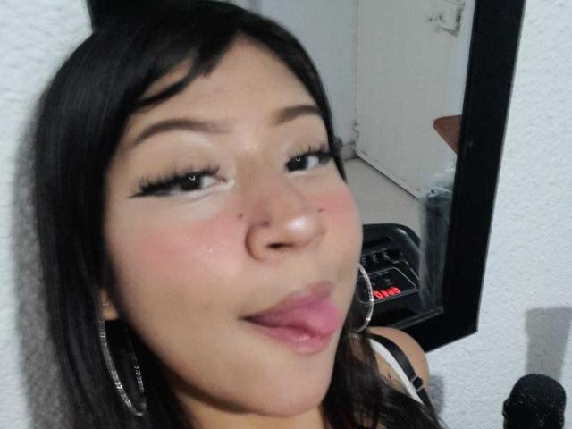 BonnieBond has dark hair, green eyes, and a pinkish blush on her cheeks. She's sticking out her tongue and wearing large hoop earrings. Her makeup includes long eyelashes.