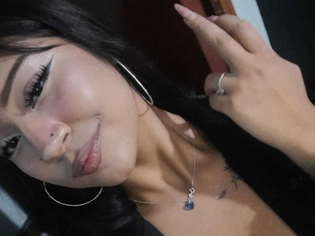 BonnieBond has long black hair, wearing large hoop earrings, a silver necklace with a blue gem, and a ring. She's smiling with a subtle nose ring and tattoo.