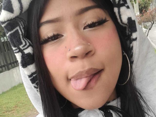 BonnieBond with black hair, hoop earrings, nose piercings, and tongue out, wearing a black-and-white hooded shirt, has her eyes half-closed and long eyelashes.