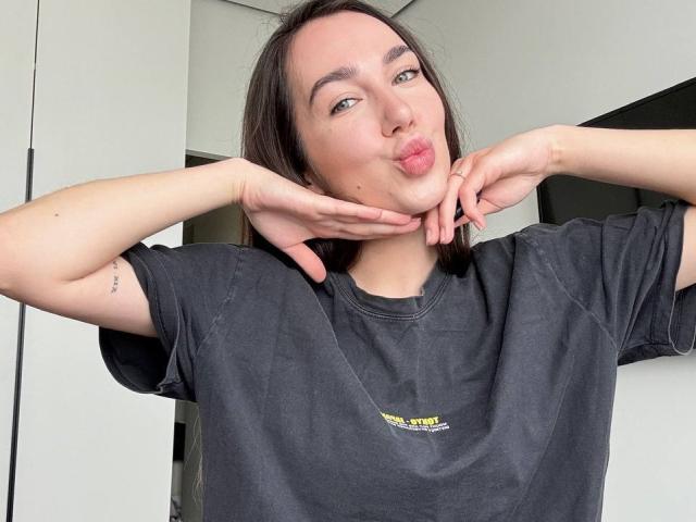 AraiaSheri puffs out her cheeks, hands under her chin, wearing a black tee, with a small yellow and green logo, in a white-walled room.