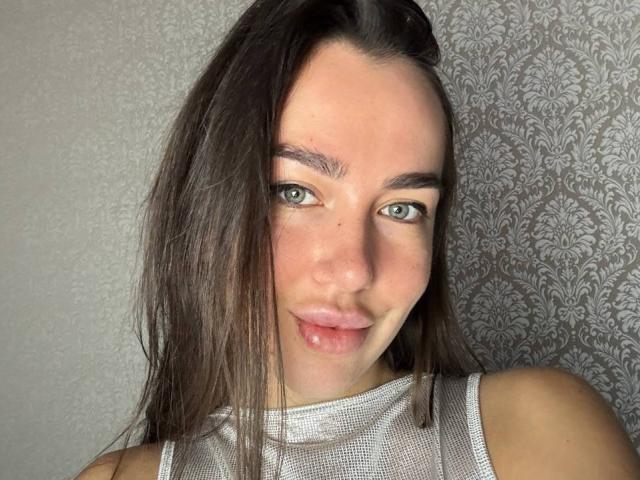AraiaSheri has green eyes, brown hair, and is pouting with slightly parted pink lips. She's wearing a shiny silver sleeveless top. Her face is slightly dirty.