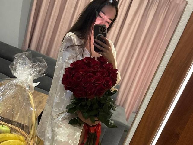 AraiaSheri holding a massive bouquet of red roses, wearing a white floral dress, reflected in a mirror, with a pink curtain and a basket of bananas in background.