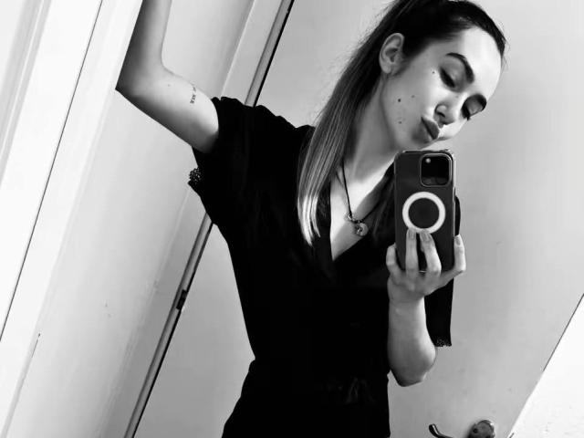 Black-and-white selfie of AraiaSheri with long hair in a ponytail, wearing a black shirt, holding a phone with a ring, puckering lips, tattoo on upper arm, small moles.