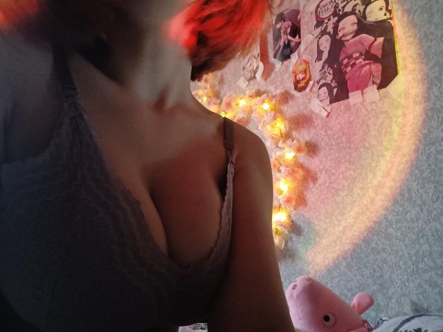 MireilleSpotwood with red hair, wearing a lace bra, showing her tits, against a wall with anime posters and glowing lights. Pink stuffed animal in the bottom right.