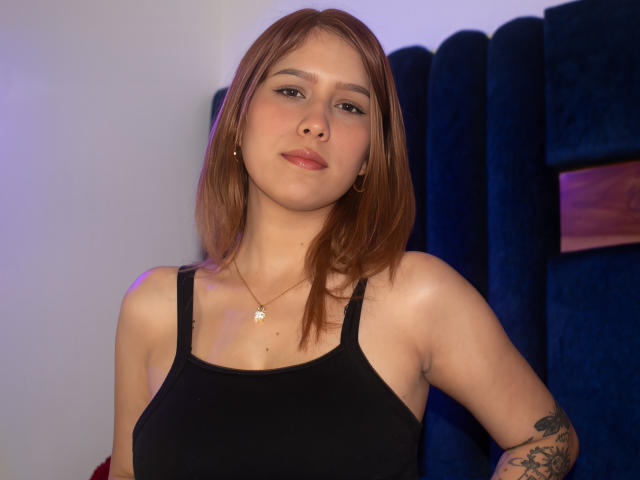 Sophie Saintclair, com cabelo castanho, vestindo uma regata preta, tem seios pequenos e uma tatuagem no braço direito, aparentando estar um pouco entediada, e está em pé em frente a uma cadeira de veludo azul..